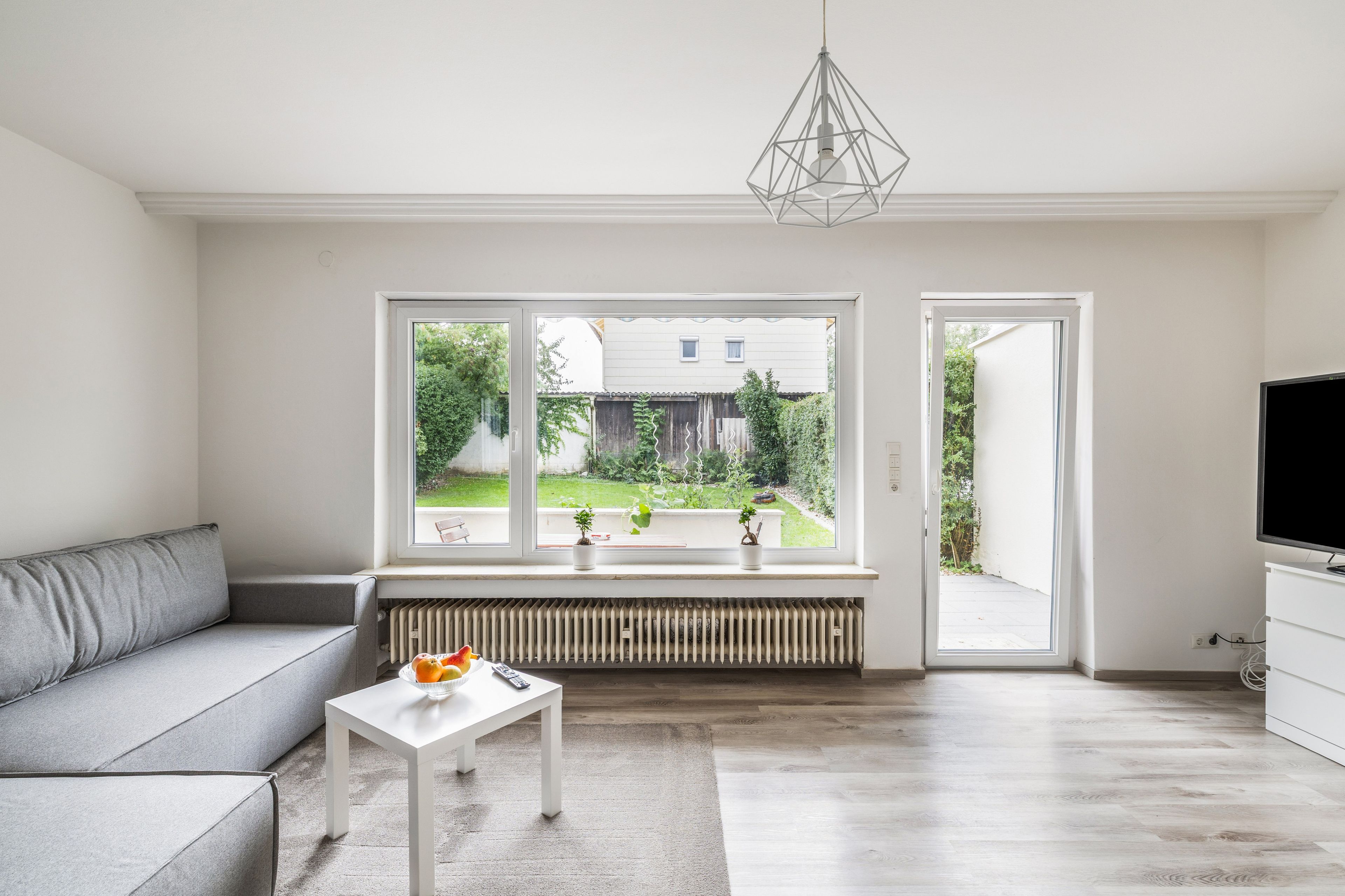 A modern living room with a grey sofa, a white table with fruit, a radiator, and a view of a garden through large windows.