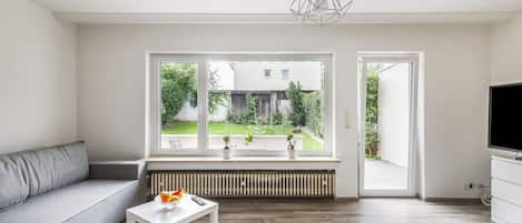 A modern living room with a grey sofa, a white table with fruit, a radiator, and a view of a garden through large windows.