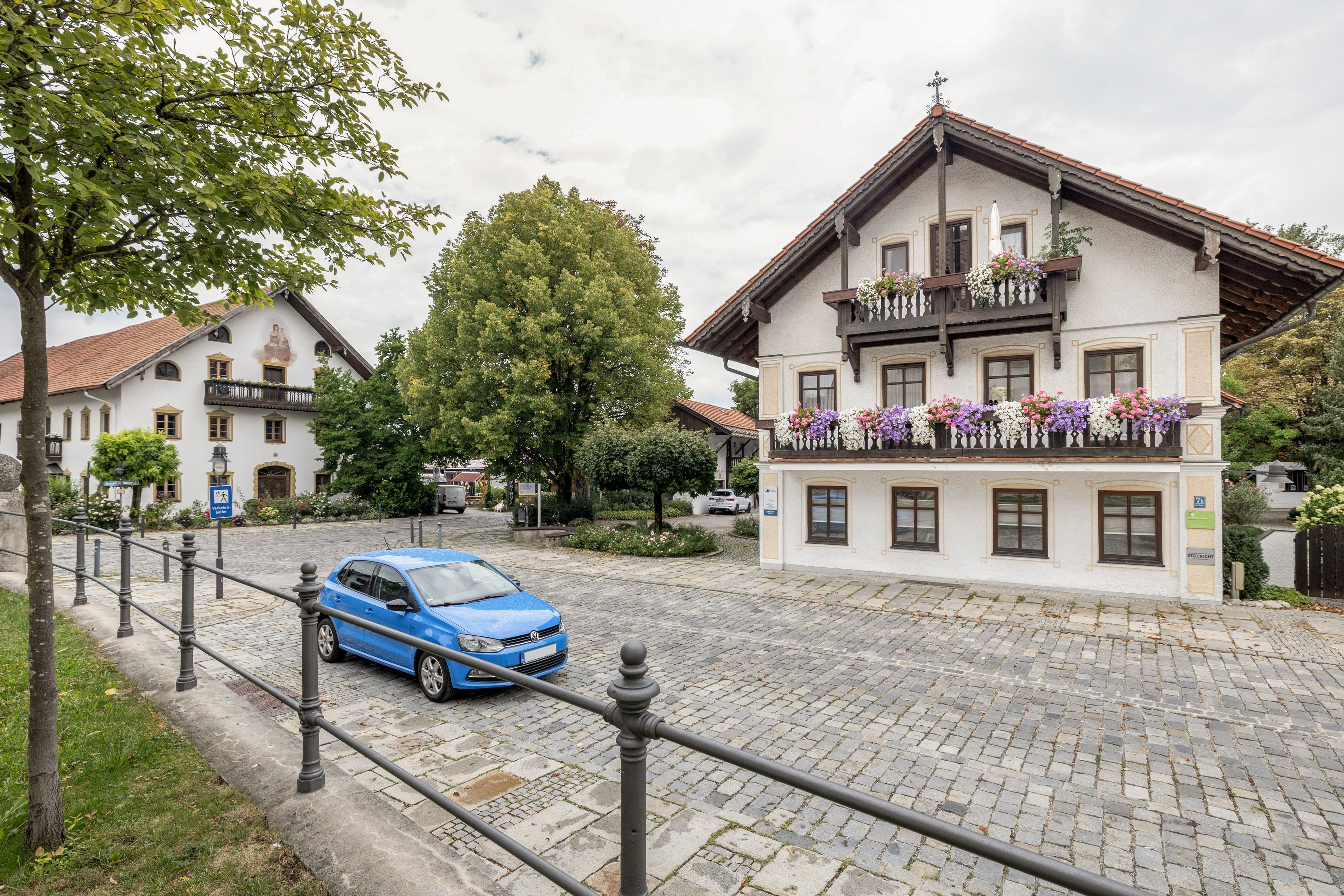 Eine Kopfsteinpflasterstraße mit einem geparkten blauen Auto am Rand, ein weißes Gebäude mit einem Balkon, der mit Blumen geschmückt ist, und ein Baum im Vordergrund.