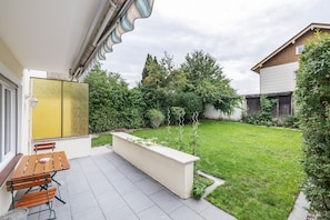 A patio with a wooden bench and a table, a garden with a lawn and various plants, and a building with a distinctive yellow and white facade. - Holiday Home 'AB12' with Private Terrace, Garden, and Wi-Fi (Oberhaching)