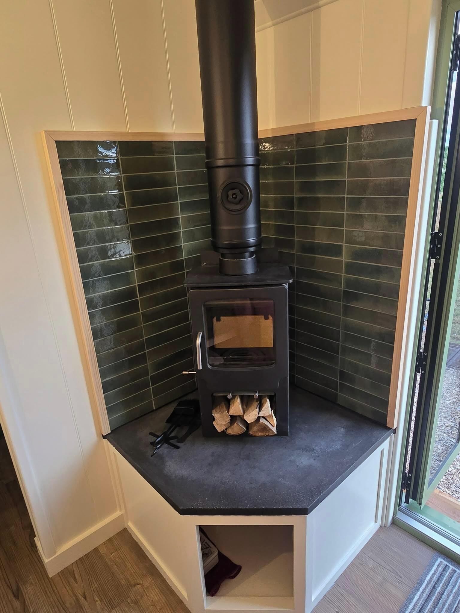 A corner room with a built-in wood stove, a small storage space below, and a view through a glass door.