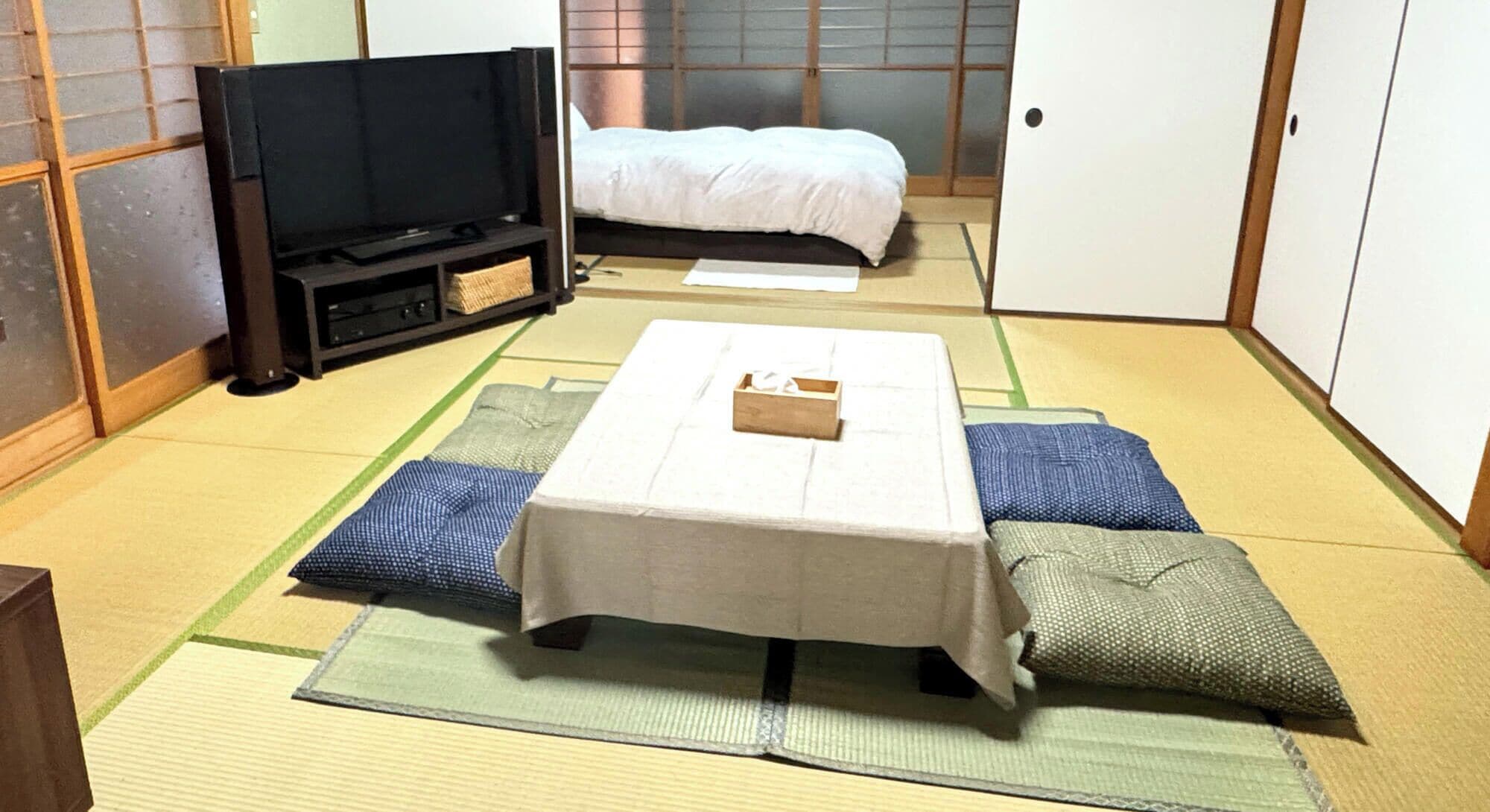 Et traditionelt japansk rum med tatamimåtter, et lavt bord med en kasse, en seng med hvidt sengetøj og et fjernsyn på et stativ.