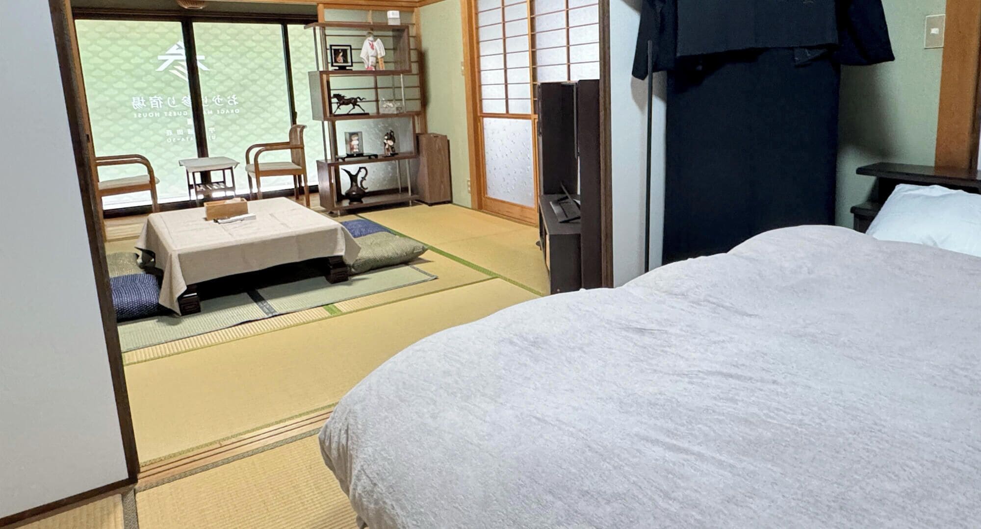 Una habitación japonesa tradicional con suelo de tatami, un futón, una pequeña mesa con una silla y una estantería con objetos decorativos.