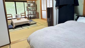Una habitación japonesa tradicional con suelo de tatami, un futón, una pequeña mesa con una silla y una estantería con objetos decorativos.