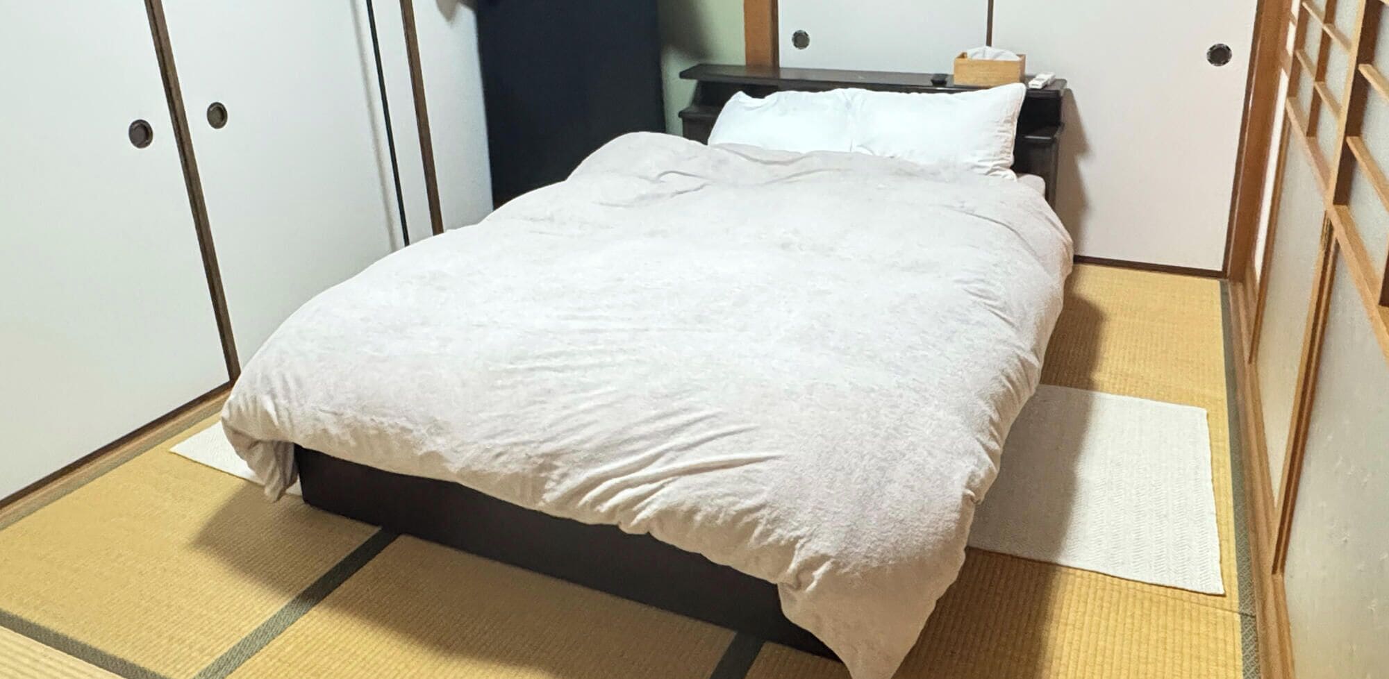 Una cama con una colcha blanca en una habitación con suelo de esteras de tatami y cabecero de madera.