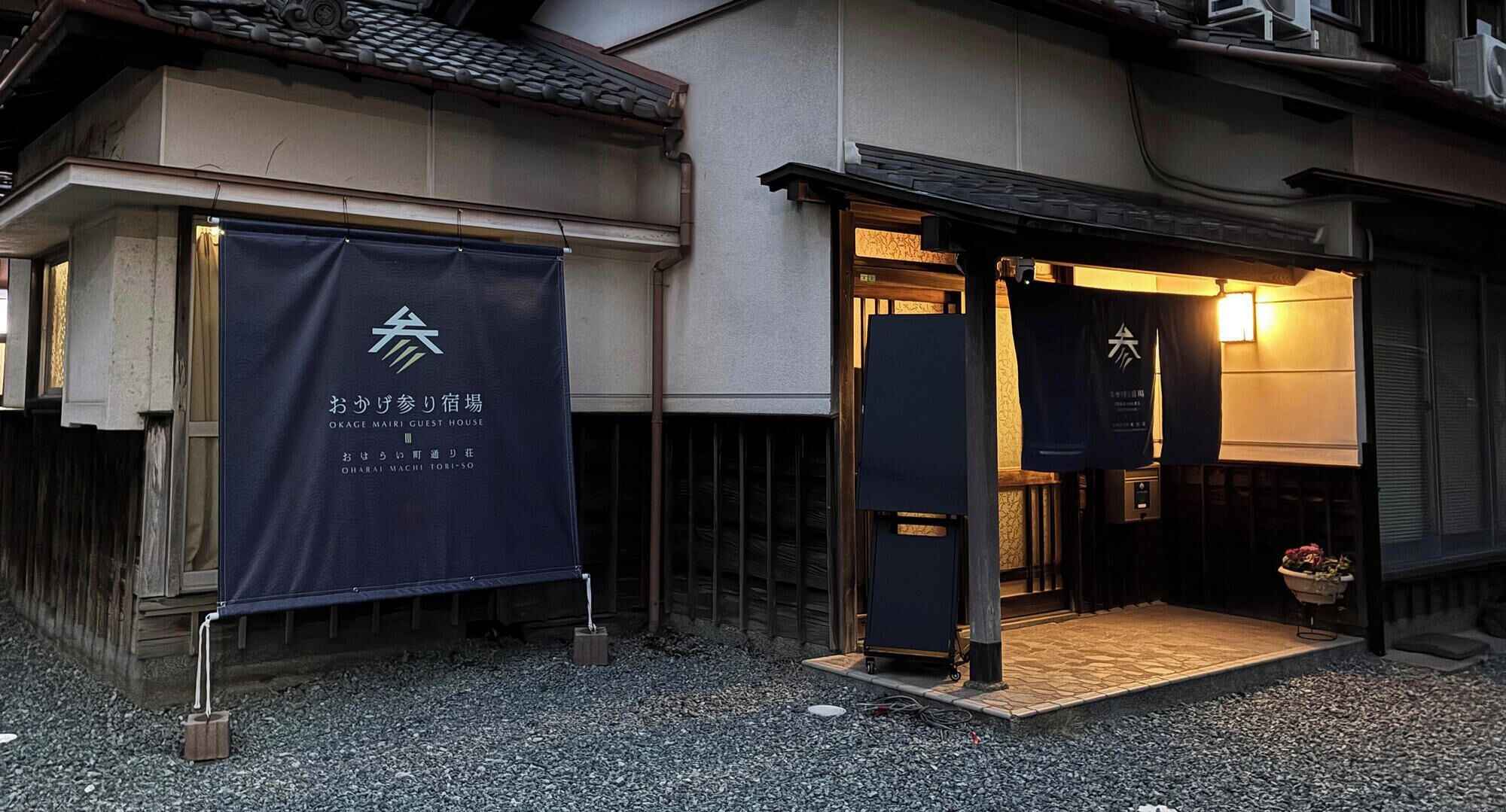 A guesthouse entrance with a dark blue banner displaying the name 'OKAGE MAIRI GUEST HOUSE' and a small entrance with a wooden mat.