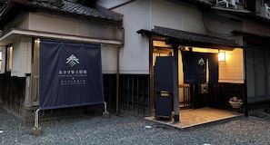 Entrada a una casa de huéspedes con un estandarte azul oscuro que muestra el nombre 'OKAGE MAIRI GUEST HOUSE' y una pequeña puerta de acceso con felpudo de madera.