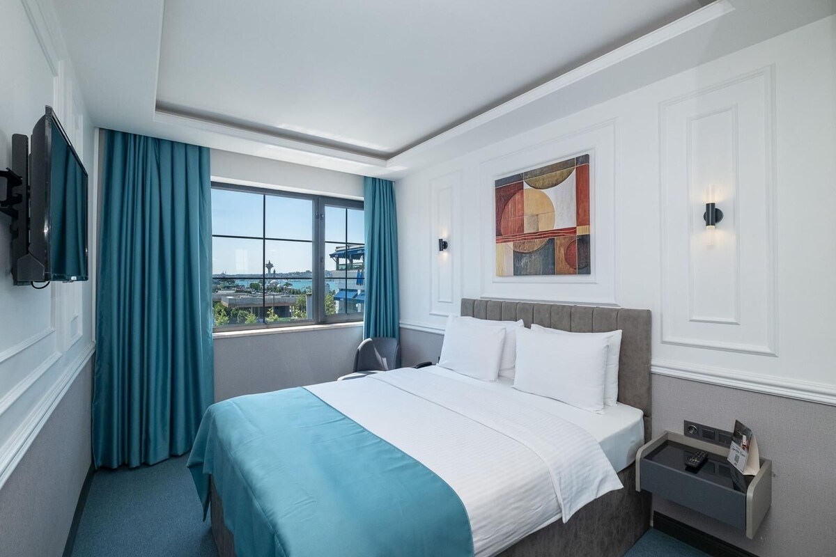 Habitación de hotel con cama, escritorio, silla, ventana con cortinas, un cuadro en la pared y mesita de noche con lámpara.