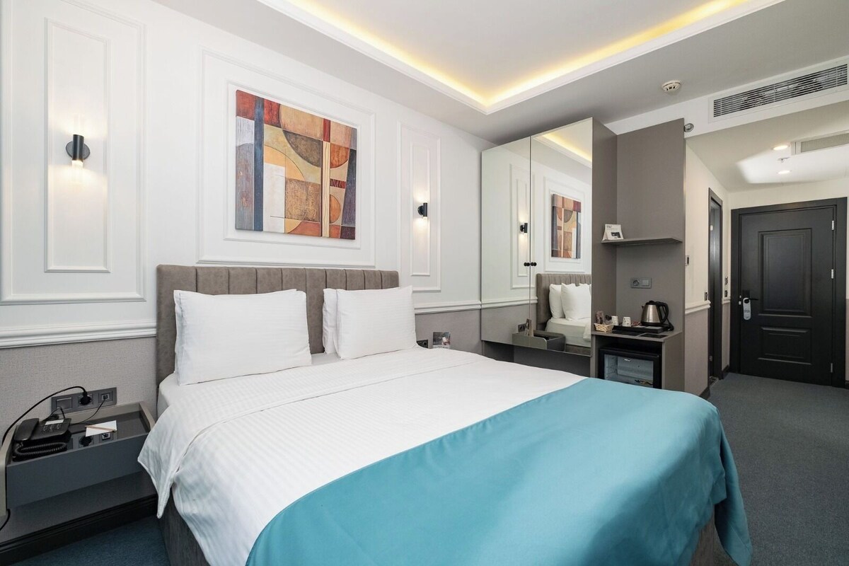 Una habitación de hotel con cama, mesita de noche, lámpara y un espejo grande.