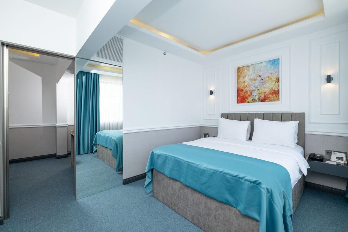 Habitación de hotel con una cama grande, una cama más pequeña, un cuadro en la pared y una mesita de noche con lámpara.