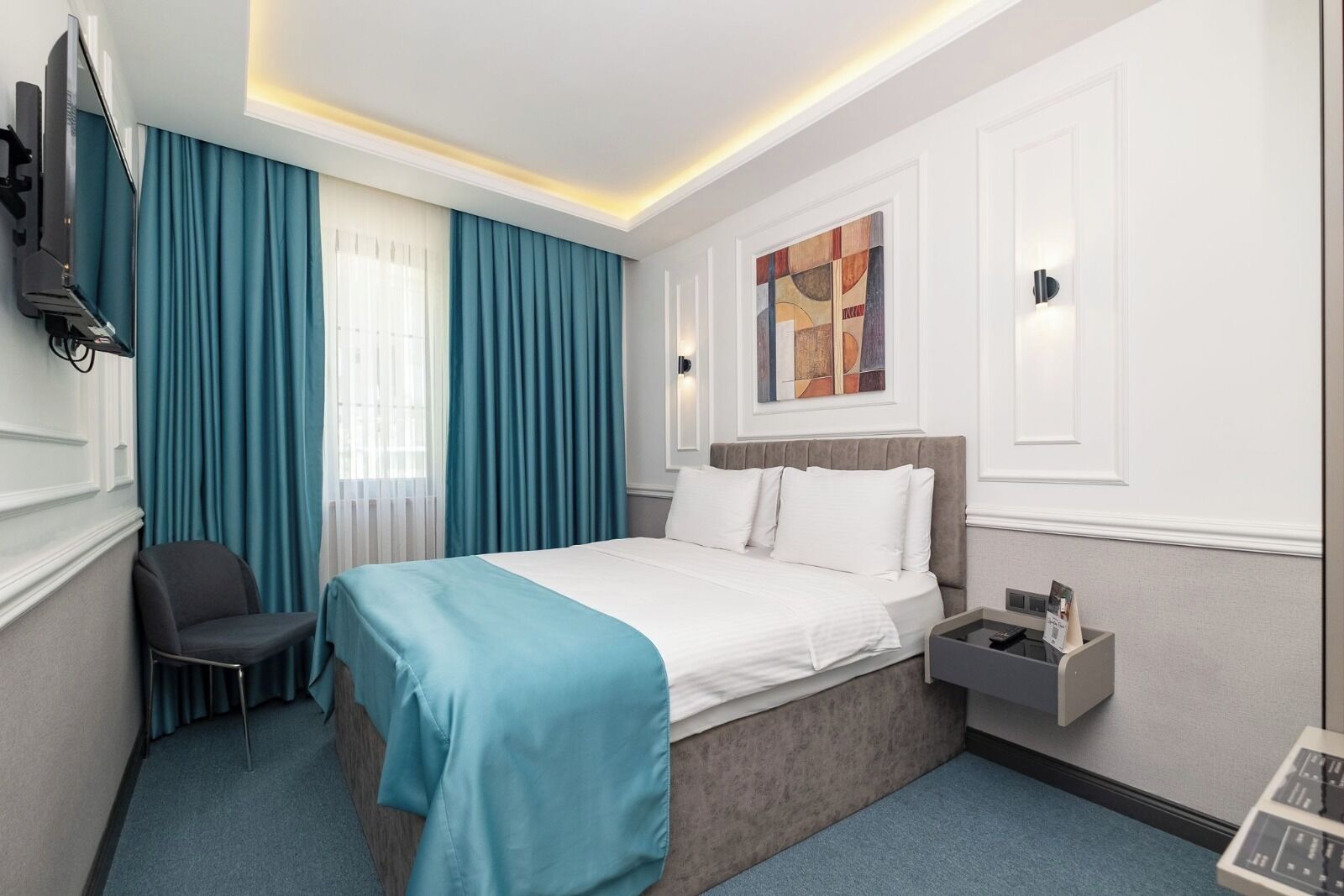 A hotel room with a bed, a chair, a desk with a lamp, and a painting on the wall.