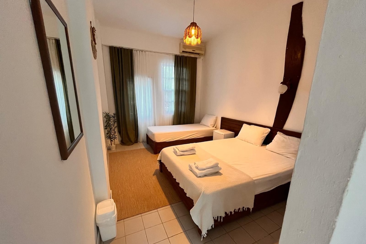 A hotel room with two beds, a bedside table, a lamp, a mirror, and a window with curtains.