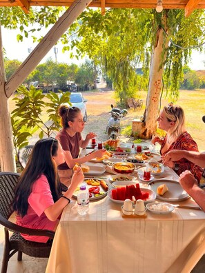 A group of people dining outdoors at a table with various dishes, under a wooden pergola with trees in the background.