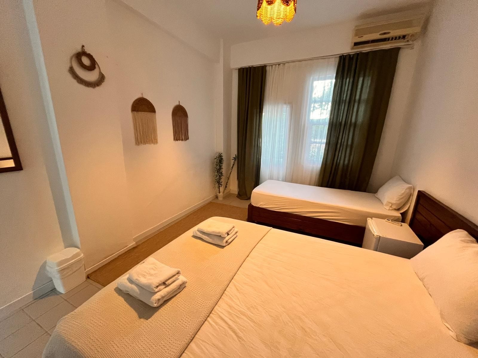 A hotel room with a bed, a bedside table, a nightstand, a window with curtains, a wall-mounted air conditioner, and a small potted plant.