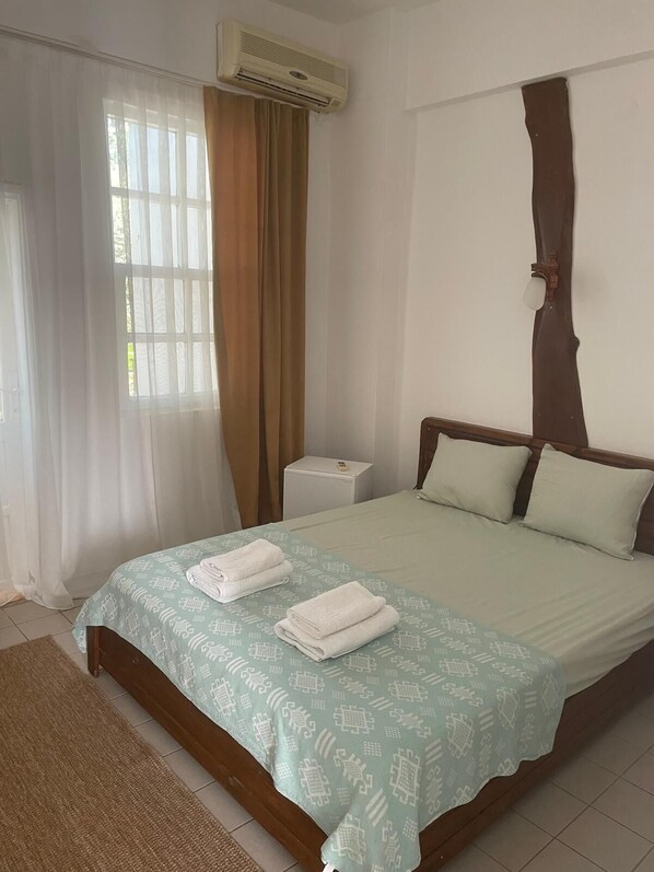 A hotel room with a bed, a bedside table, an air conditioning unit, and a window with curtains.