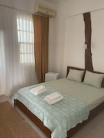 A hotel room with a bed, a bedside table, an air conditioning unit, and a window with curtains.