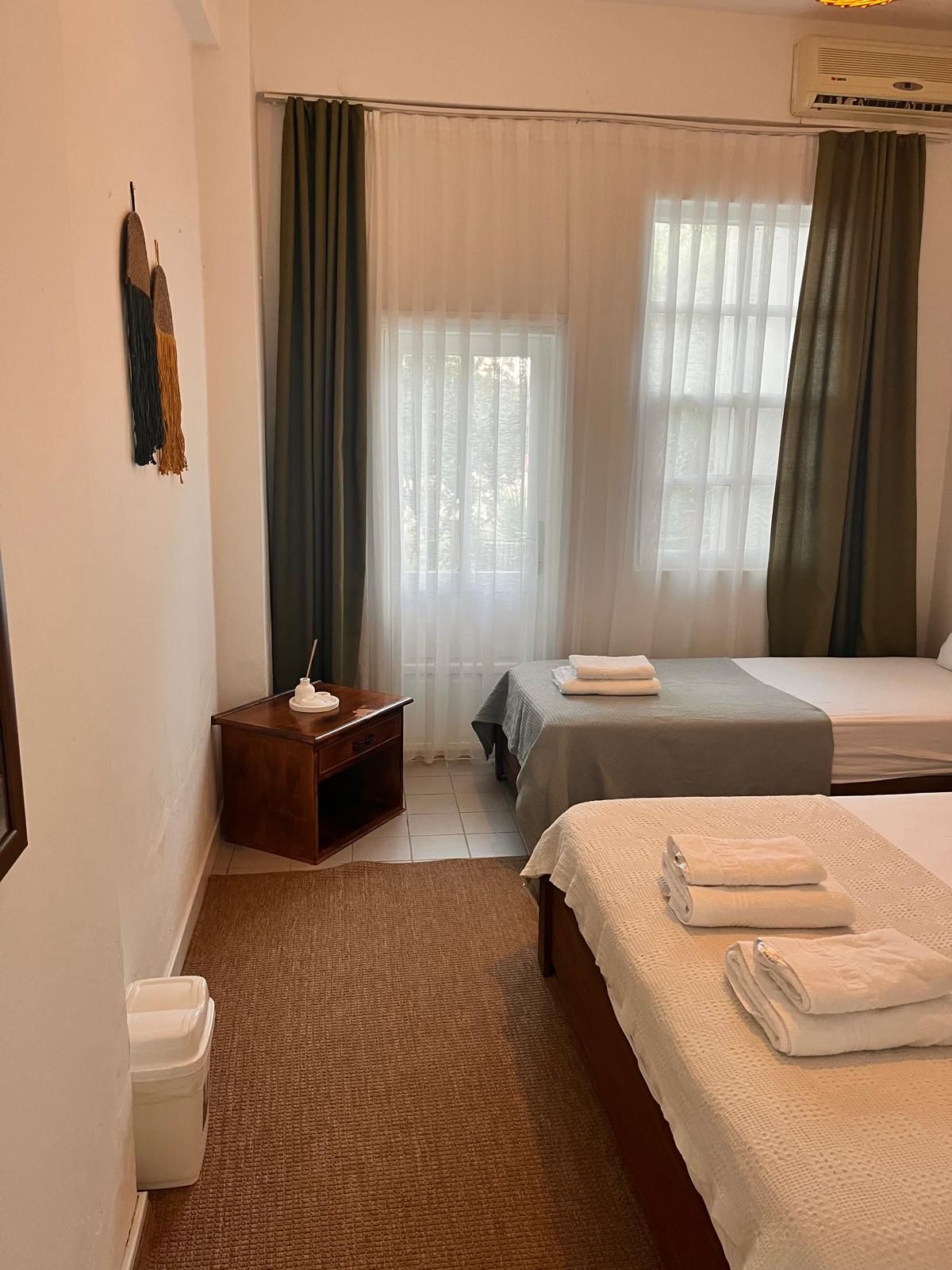 A hotel room with two beds, a small wooden nightstand, a white trash bin, and a window with sheer curtains.