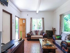 Sala de estar com sofá, mesa de centro e televisão. Uma porta dá acesso a outro cômodo.