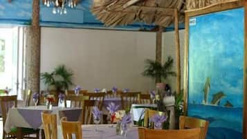 Un restaurante con mesas y sillas de madera, techo de paja y un mural de delfines.