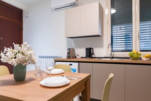 Een eethoek met een houten tafel gedekt voor twee personen, een vaas met witte bloemen en een keuken met een koffiezetapparaat en een fruitschaal.