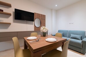 Un'area pranzo moderna con un tavolo di legno apparecchiato per due persone, una televisione a schermo piatto montata a parete, uno specchio rotondo e un divano blu sullo sfondo.