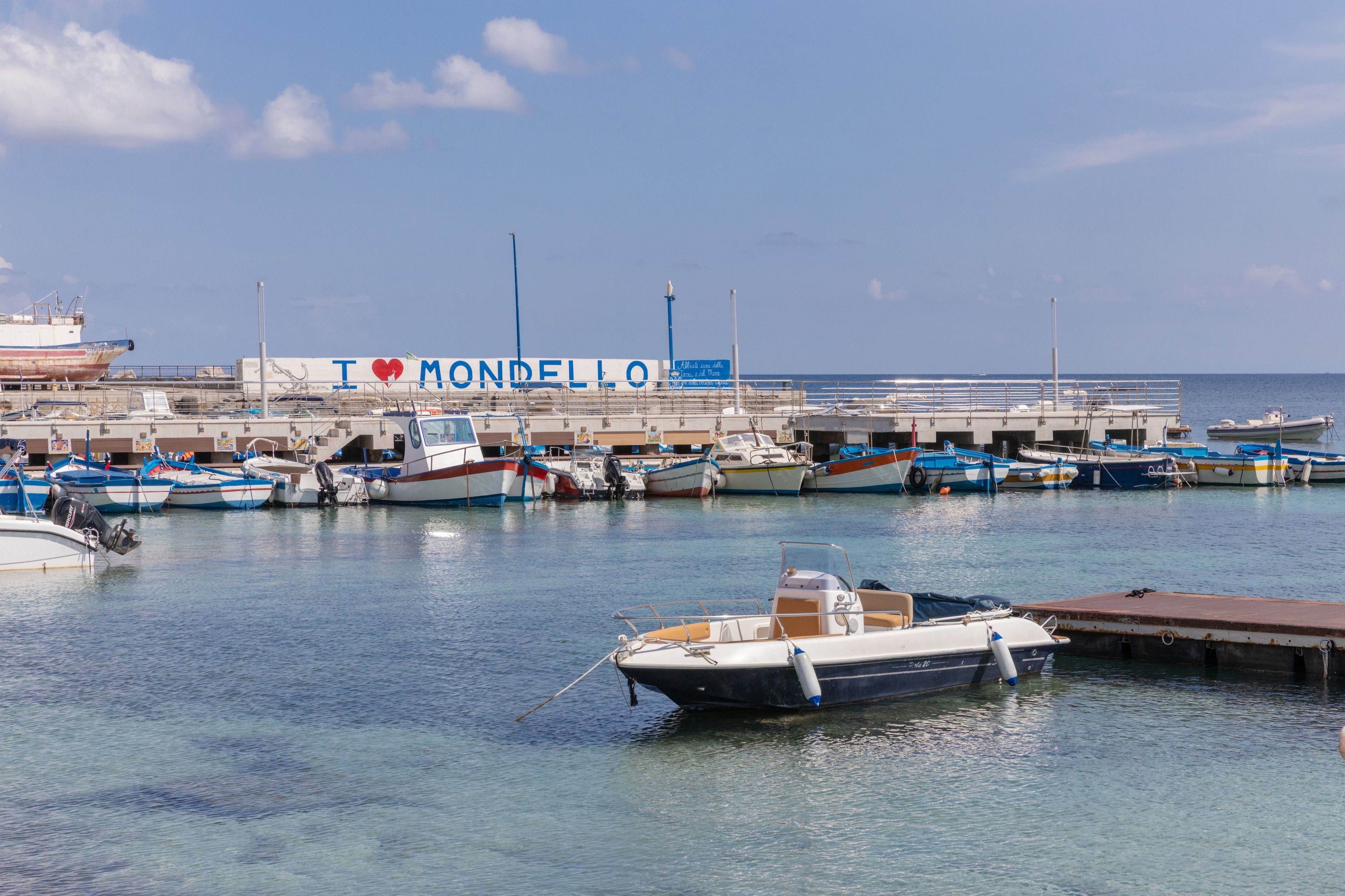Een jachthaven met aangemeerde boten, een groot gebouw met de tekst 'I ♥ MONDELLO', en een heldere lucht.