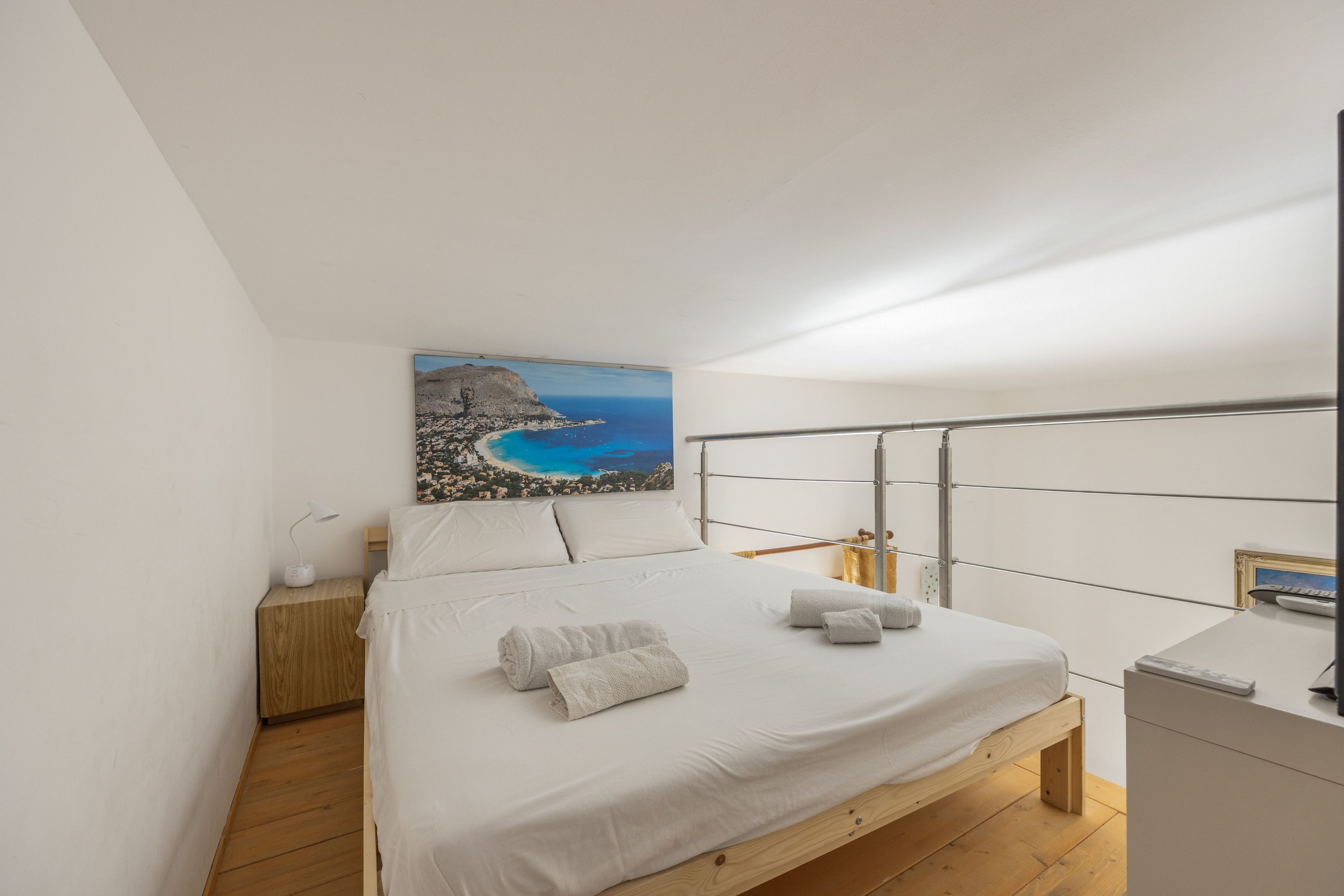 Une chambre à coucher comprenant un lit, une table de chevet, un bureau, un tableau représentant un paysage côtier et une tête de lit en métal.