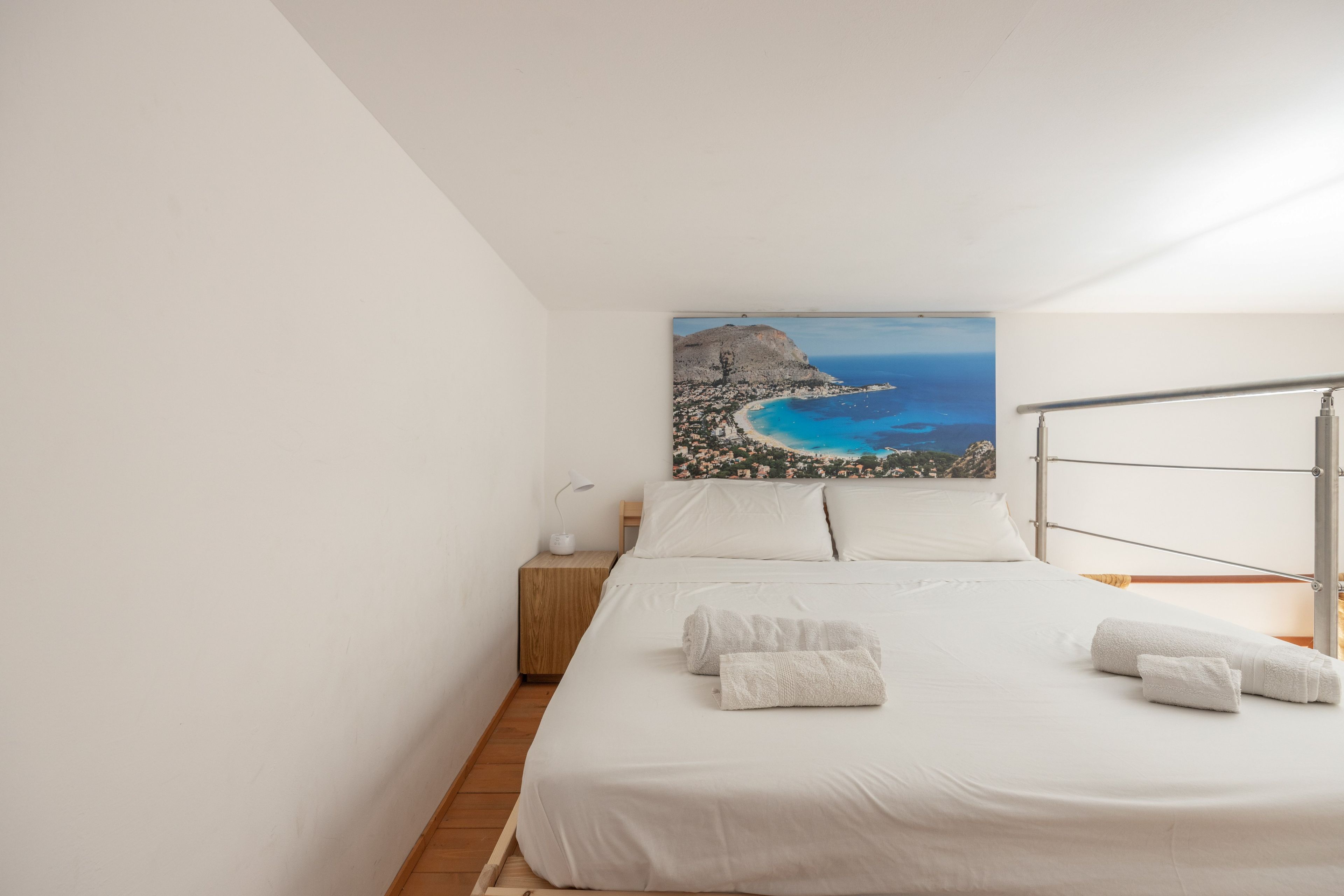 A bedroom with a bed, a nightstand, a lamp, and a painting of a coastal landscape.