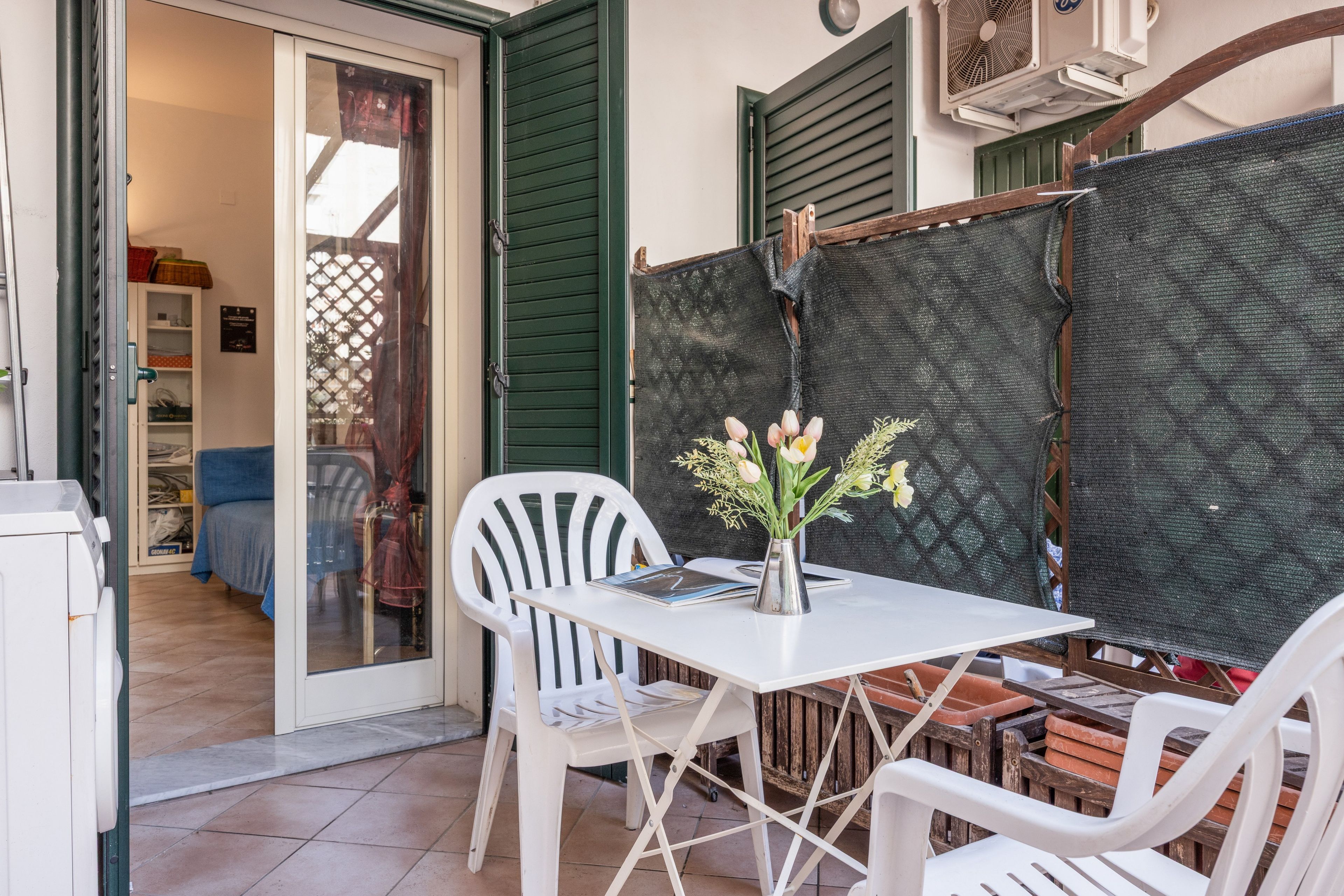 A patio area with a table and chairs, a small vase with flowers, and a view of the interior through a glass door.
