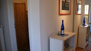 Een hal met een houten deur, een witte plank met een fles, en lijstjes met foto's aan de muur.