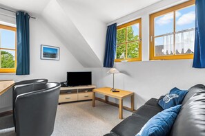 A modern room with a sloped ceiling, a flat-screen TV, a small table with a lamp, a sofa with blue cushions, and a picture on the wall. - Modern beachfront apartment with exceptional sea views at Schönberger Strand (Schönberg (Holstein))