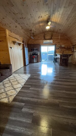 An interior with a wooden ceiling, a kitchen area with appliances, a dining area with chairs, and a large window.
