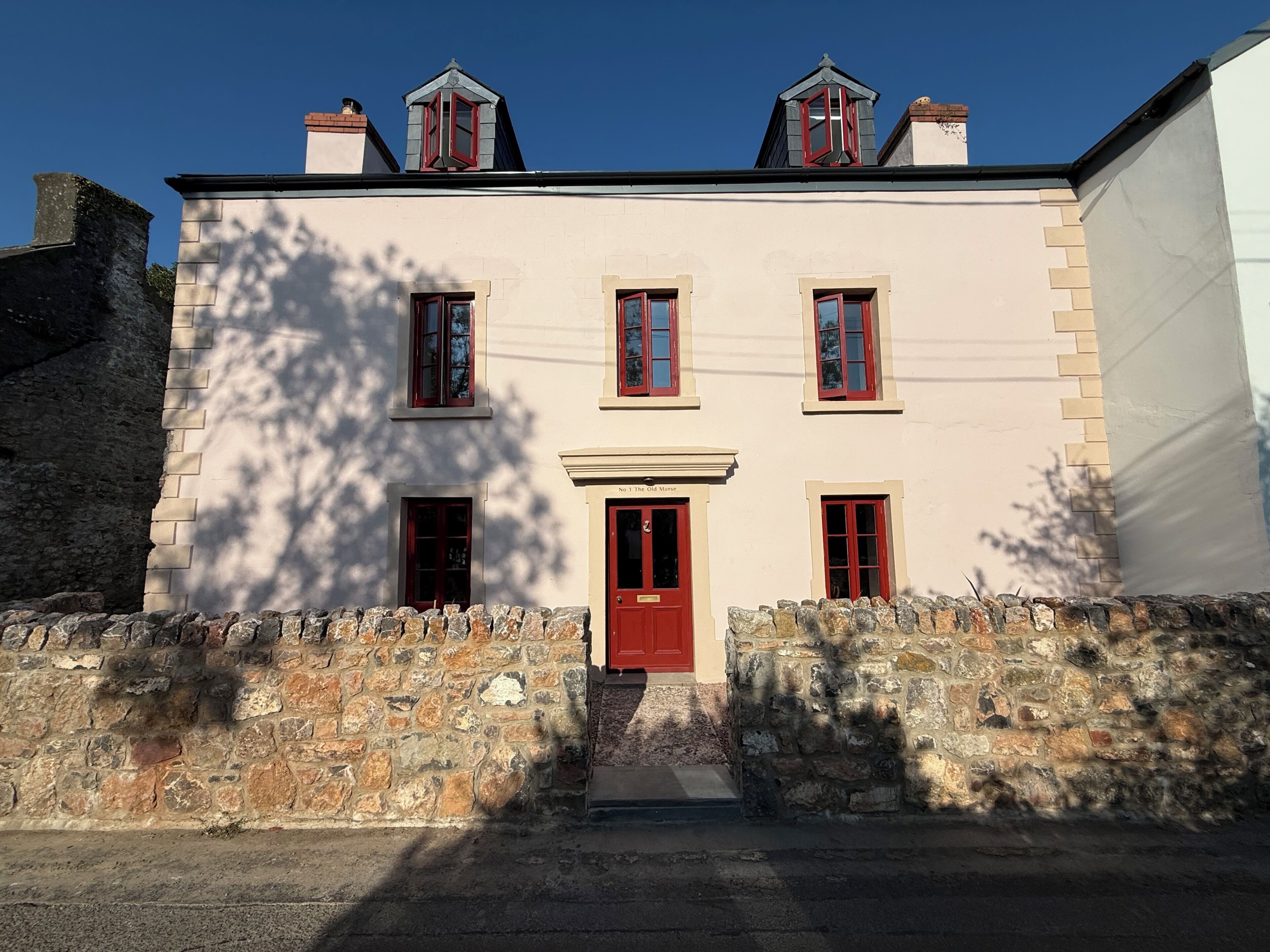Edificio de dos pisos con una puerta roja y varias ventanas, rodeado por un muro de piedra.