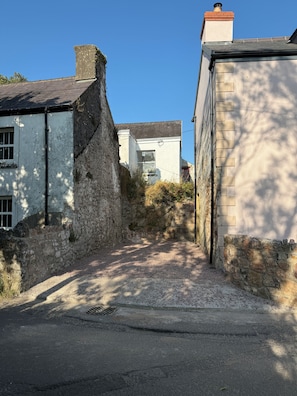 A narrow street with stone walls and a paved path leading to a white building with a visible window.