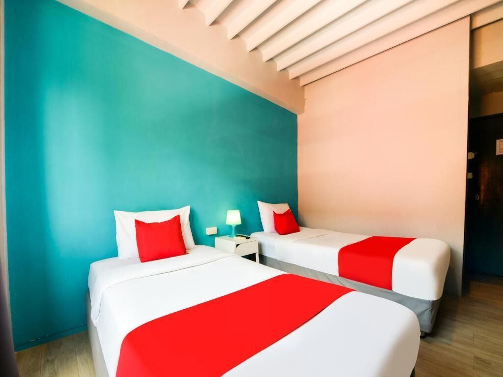 A hotel room with two beds, each with white bedding and red accents, a nightstand with a lamp, and teal and peach walls.