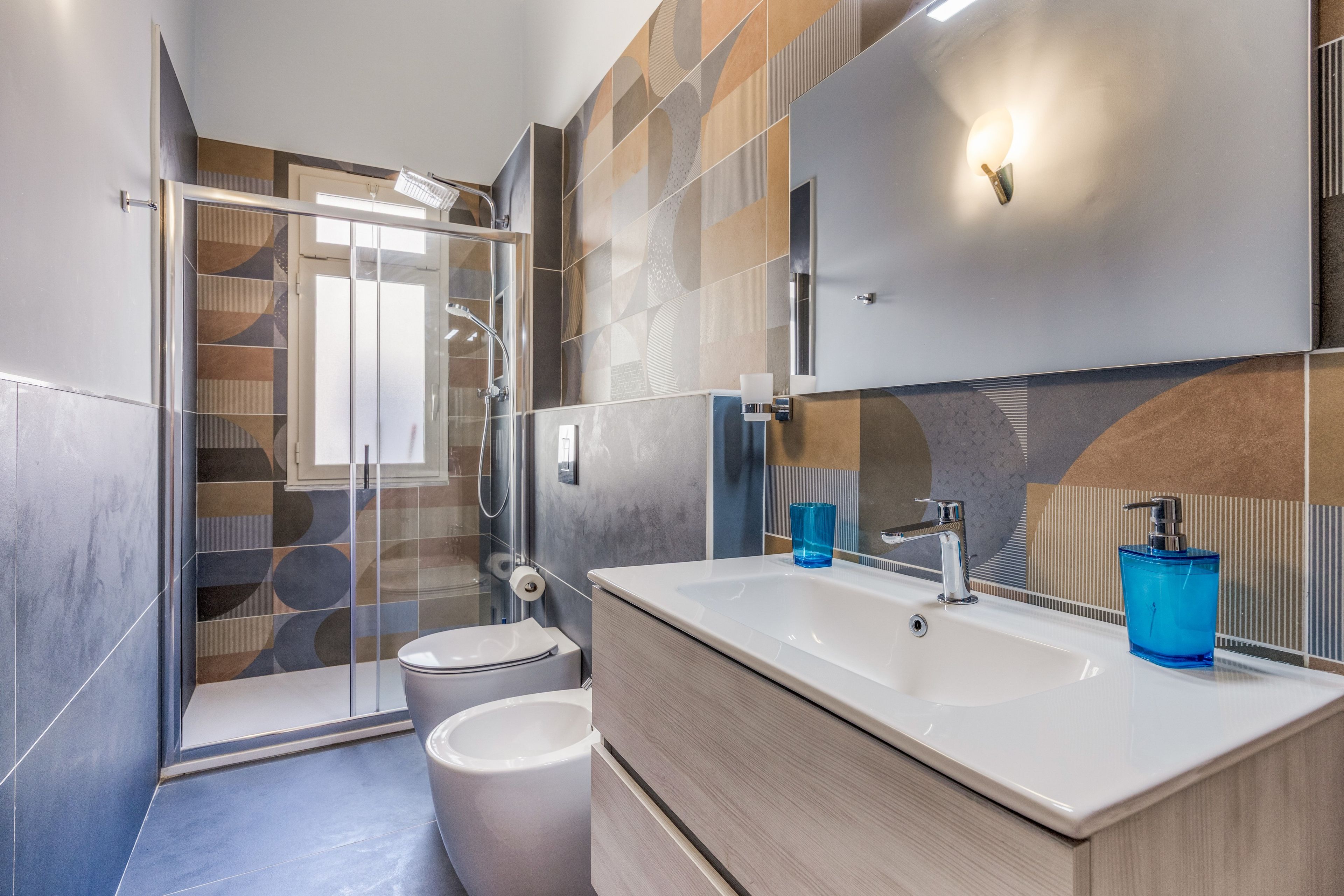 Een moderne badkamer met een glazen douchecabine, een wit toilet, een wastafel met spiegel en een blauwe zeepdispenser.