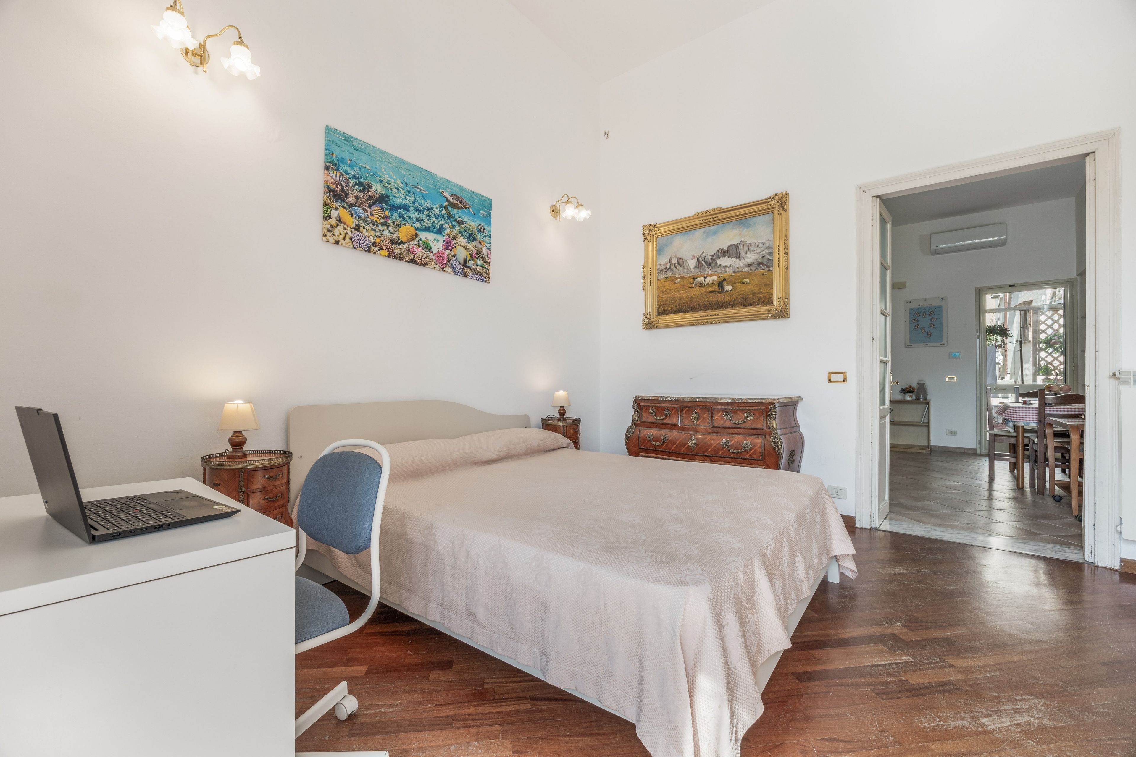 Een slaapkamer met een bed, een bureau met een laptop, een stoel, een kaptafel en twee schilderijen aan de muur.