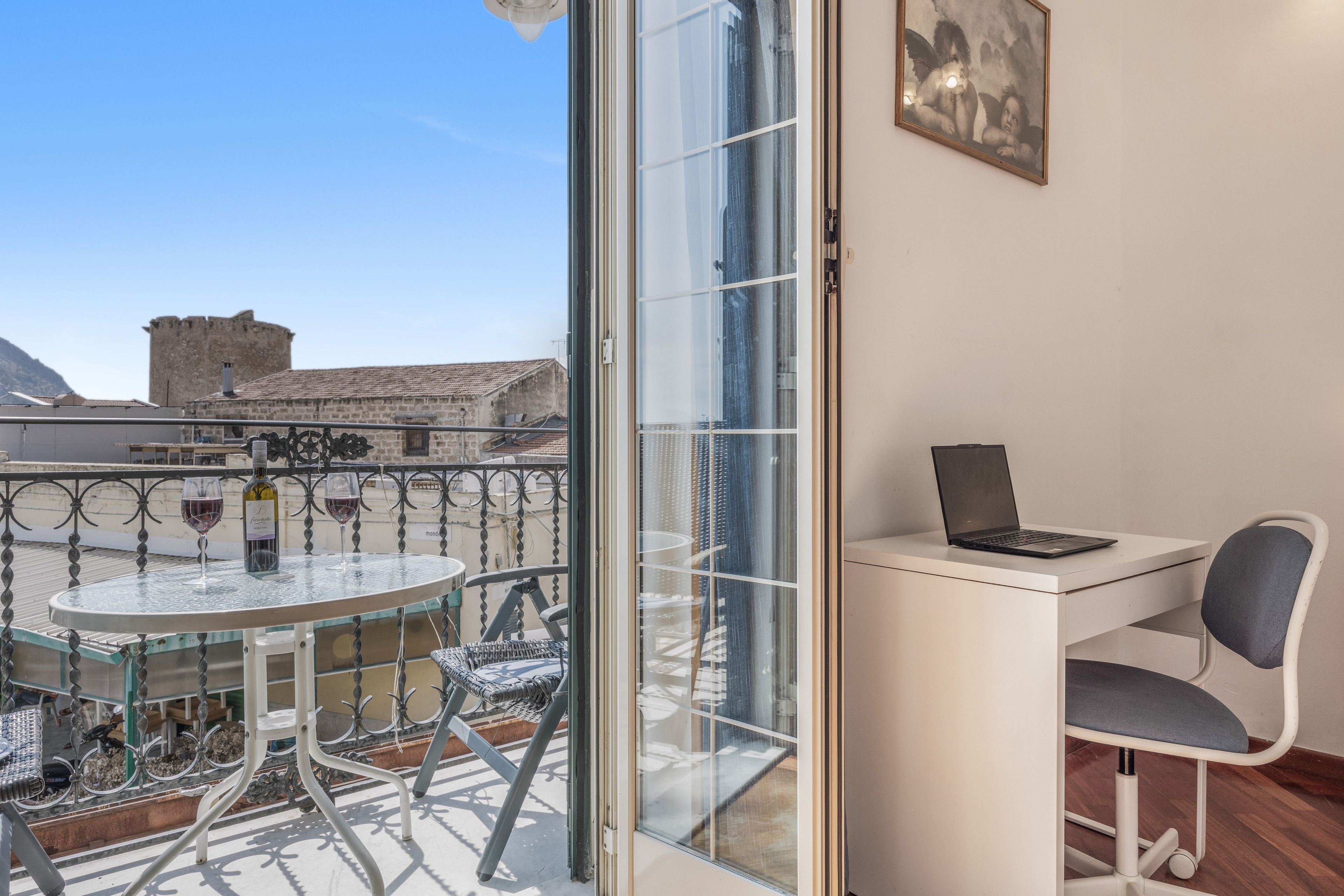 Ein Balkon mit Glastür, einem runden Tisch mit Stühlen, einem Laptop auf einem Schreibtisch und einem Sessel.