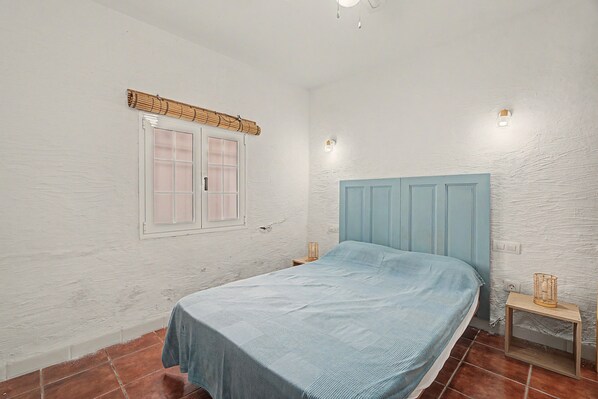 Une chambre à coucher avec un lit, une fenêtre avec des stores, une lampe fixée au mur et une petite table de chevet.