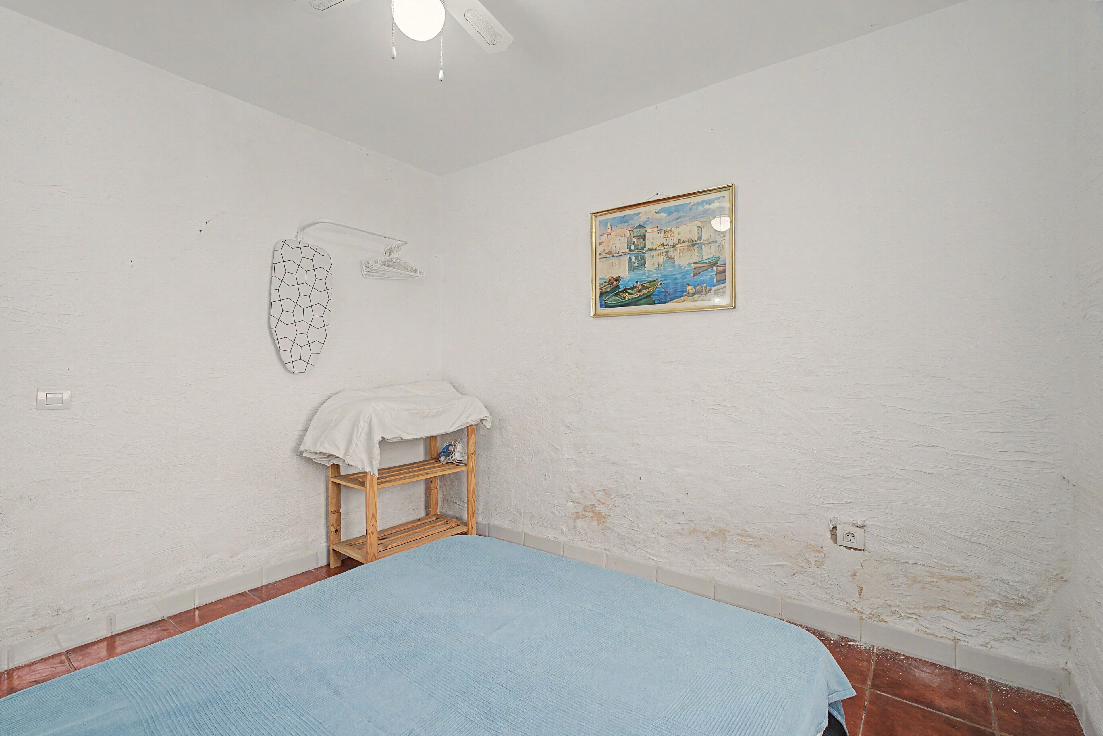 A room with a bed, a small wooden shelf, a wall-mounted light fixture, and a framed painting.
