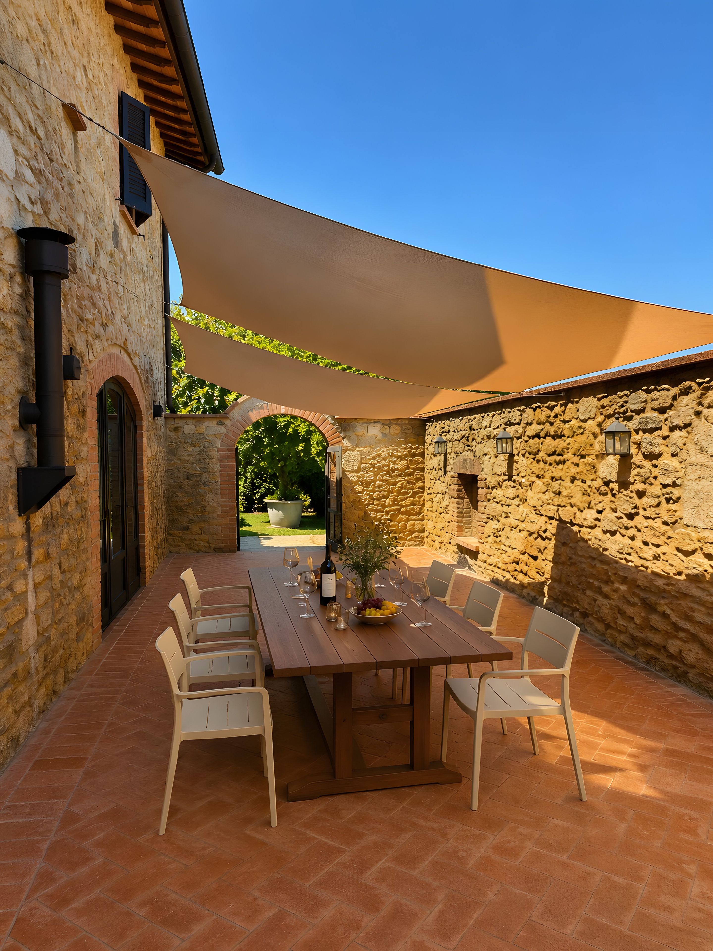An outdoor patio with a wooden table and chairs, surrounded by stone walls and a large shade sail.