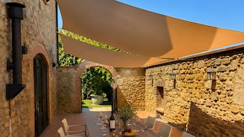 An outdoor patio with a wooden table and chairs, surrounded by stone walls and a large shade sail.