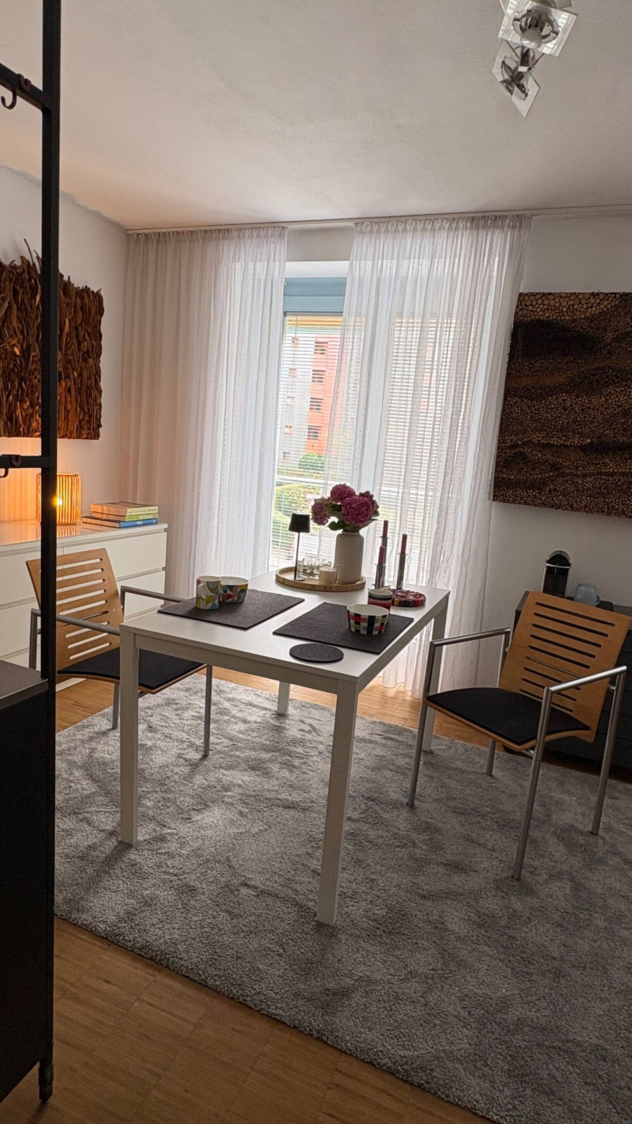 Un espace de salle à manger moderne avec une table blanche, deux chaises, un tapis et une vue sur les bâtiments à travers des rideaux transparents.