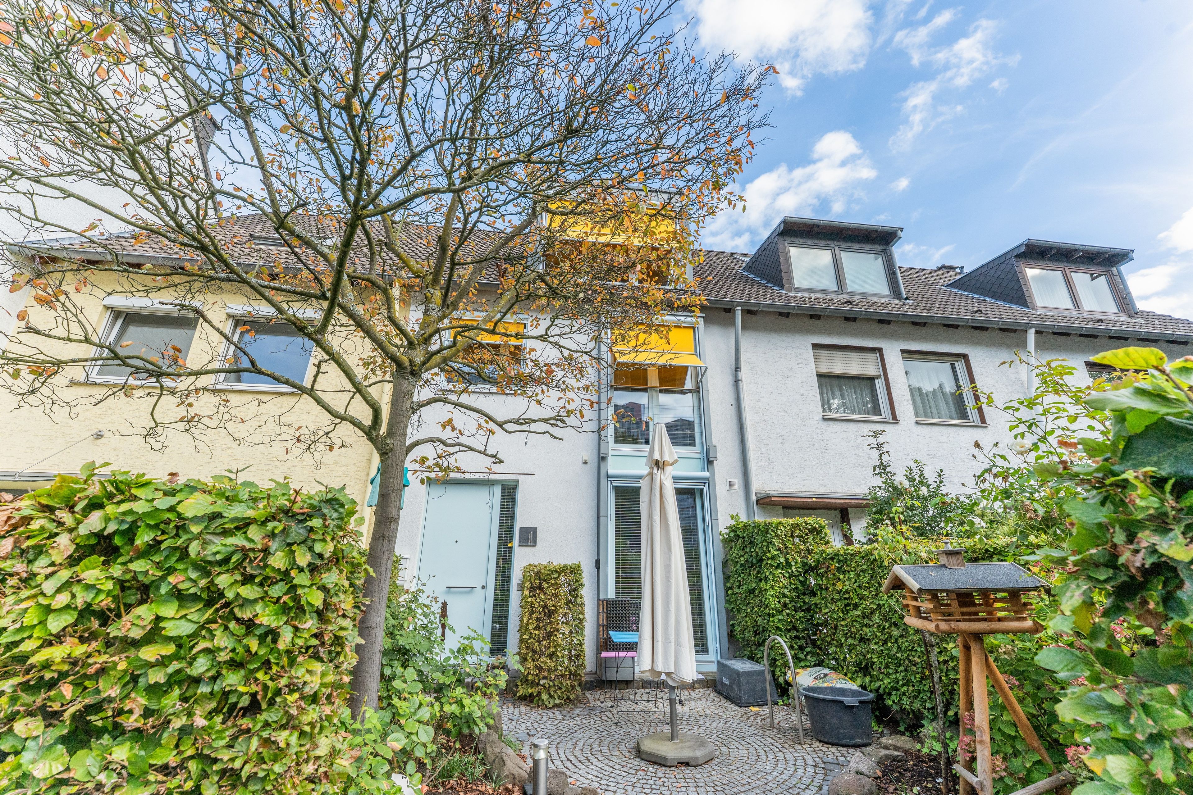Un immeuble résidentiel avec un toit recouvert de tuiles, une porte bien visible et un petit jardin agrémenté d’un arbre et de quelques arbustes.