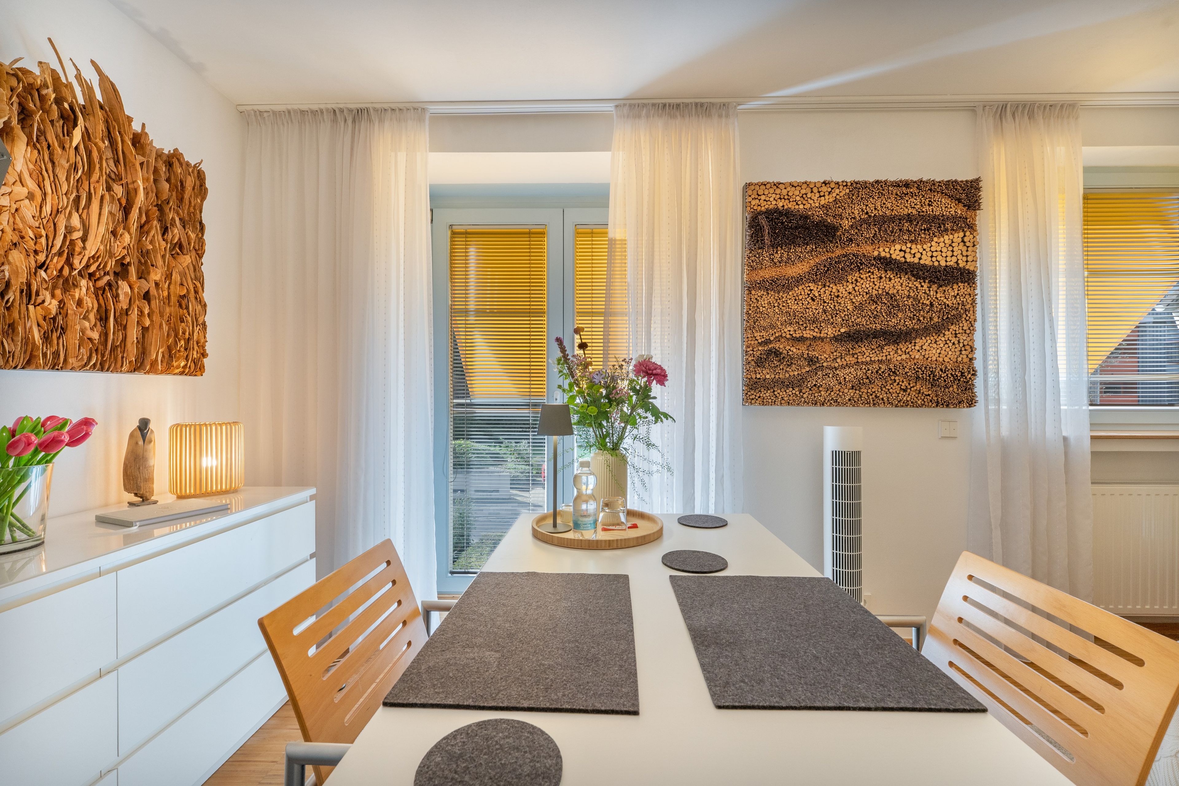 Un espace repas moderne avec une table blanche, des sets de table gris, des chaises en bois et un vase rempli de fleurs.