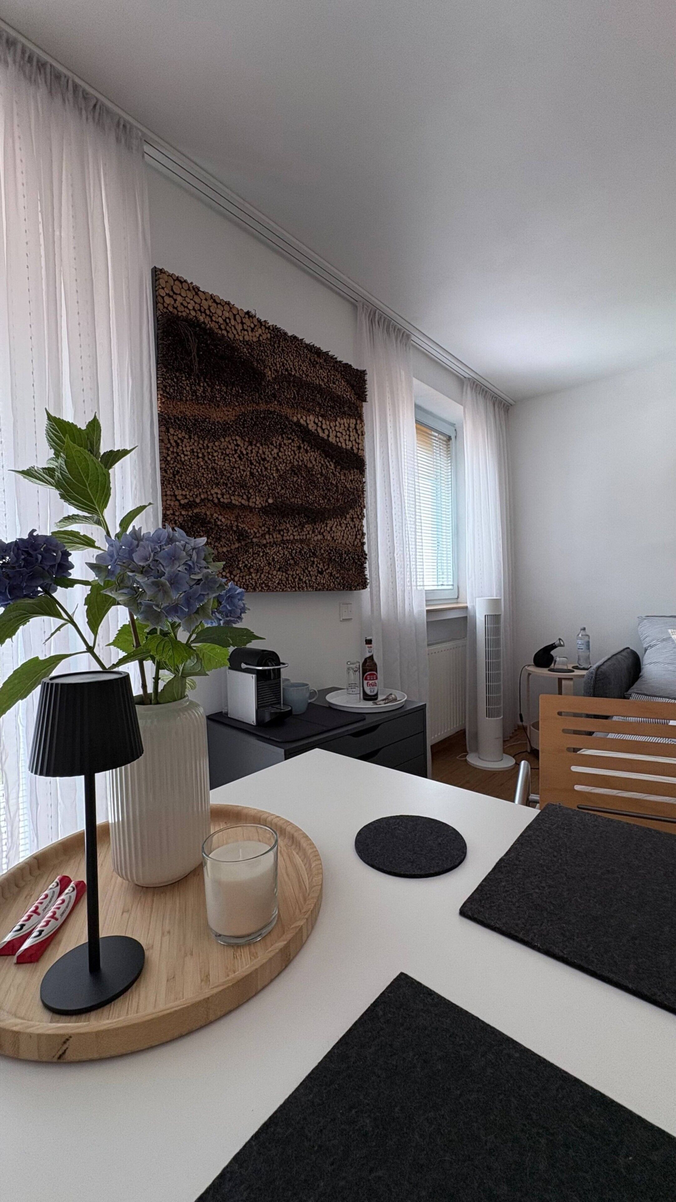Ein modernes Zimmer mit einem weißen Tisch, einer Wanddekoration, einer Topfpflanze und einem Nachttisch mit einer Lampe.