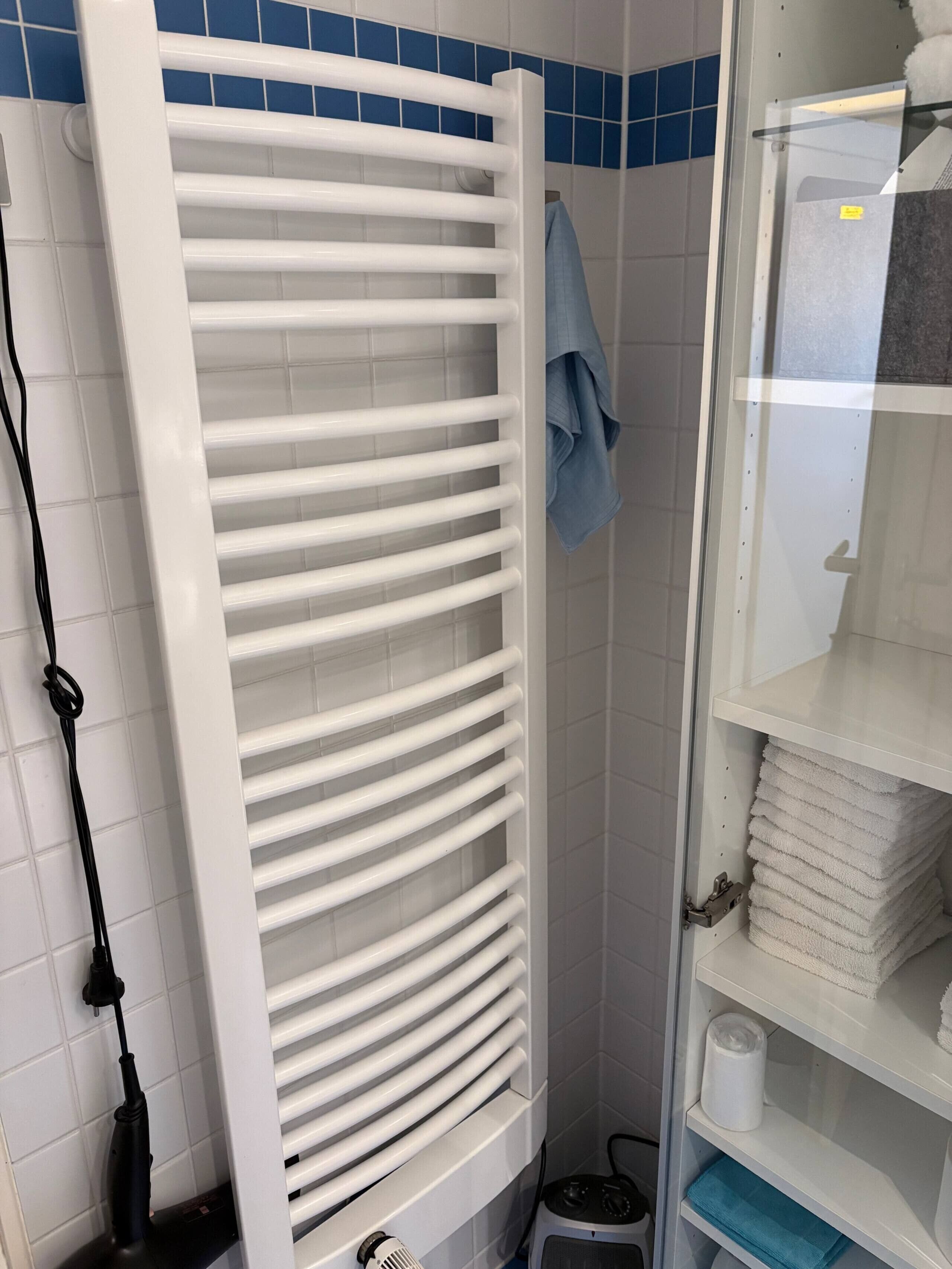 Une salle de bain avec un porte-serviettes blanc, une serviette bleue suspendue, et une étagère où sont pliées des serviettes, ainsi qu'un rouleau de papier toilette.