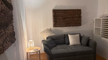 Una sala de estar con un sofá gris, una mesita redonda con una lámpara y un mueble blanco con estantes.