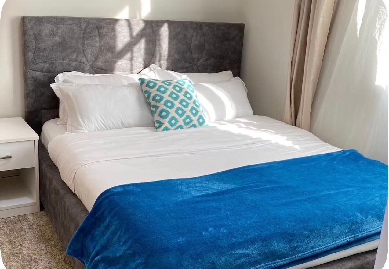 Una cama con cabecera gris, ropa de cama blanca y una manta azul.