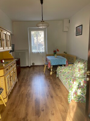 Una habitación con piso de madera, una mesa de comedor con mantel azul, un sofá con funda floral, un mueble con paneles decorativos y una ventana con vista a palmeras.